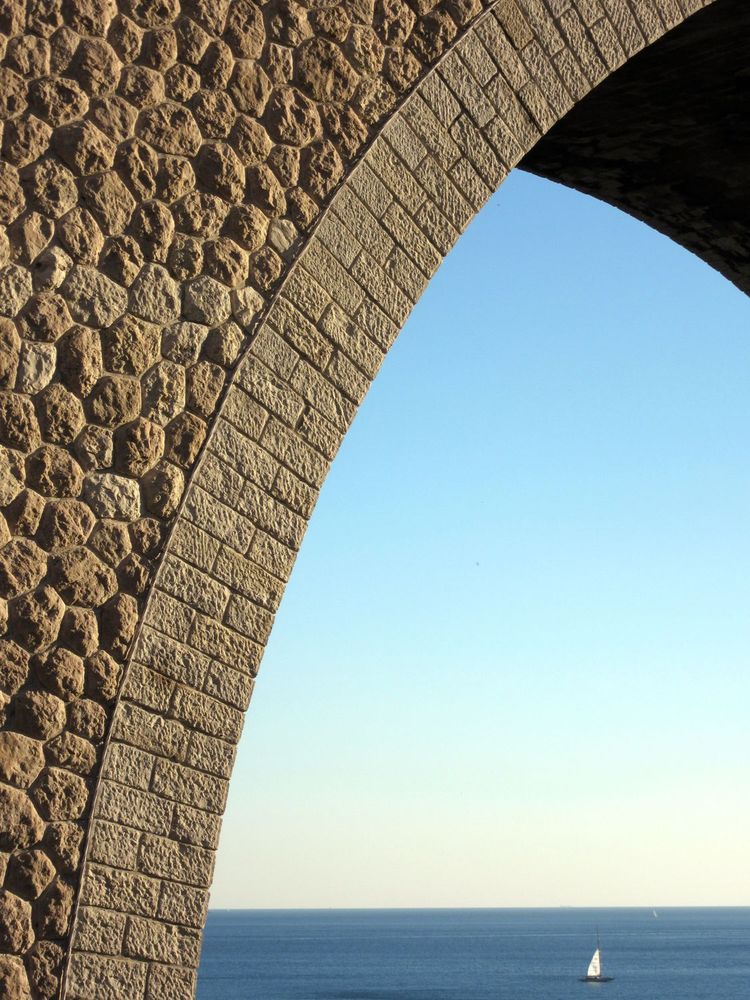 Sous une arche du viaduc de Corbières à Marseille, un voilier navigue sur une mer plate.