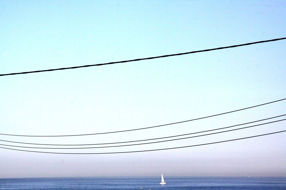Un voilier au large des Goudes à Marseille navigue sur une mer plate. Au-dessus, dans le ciel, des fils électriques.
