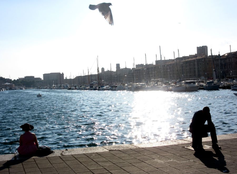 Sur le Vieux-Port de Marseille, deux personnages : un homme et une femme au chapeau. Au milieu, un pigeon vole.