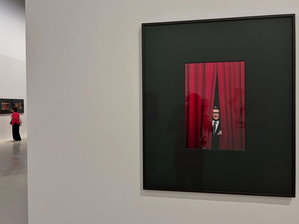 Une photo d'Yves Saint-Laurent entrouvant un rideau rouge de théâtre. Plus en retrait, sur la gauche, une femme, en rouge, regarde une photo.