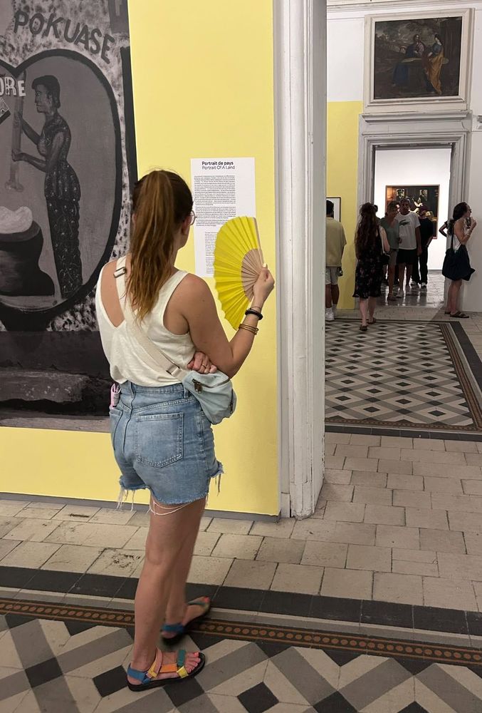 Une femme, avec un éventail jaune, lit une fiche plaquée sur un mur jaune aux Rencontres de la photographie à Arles.